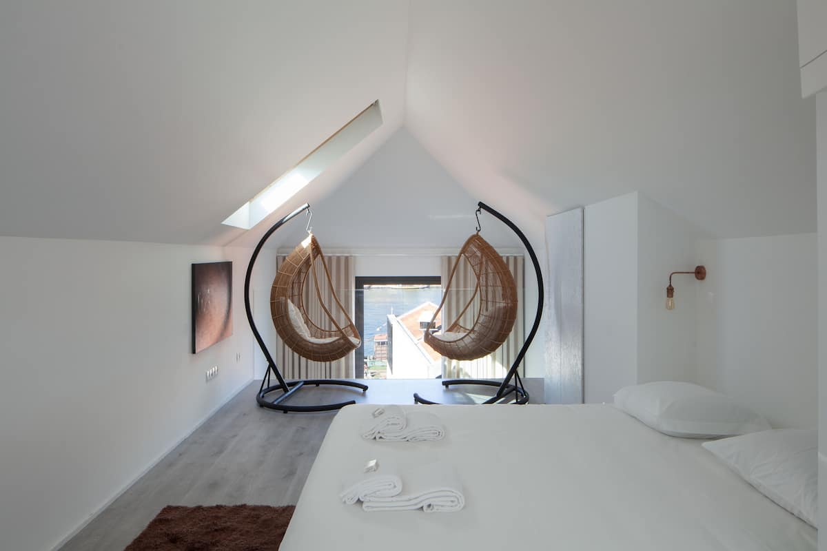 A modern bedroom with a bed, two hanging chairs, a view of the sea, and a skylight.