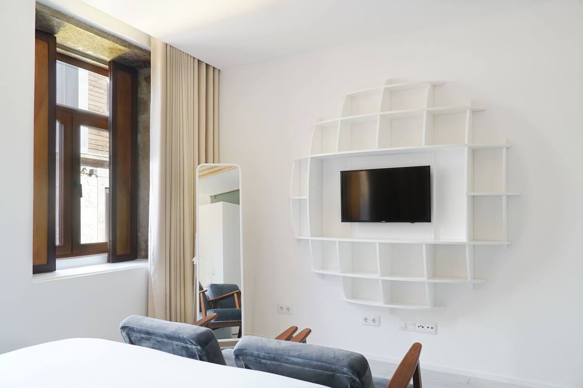 A hotel room with a large window, a flat-screen TV mounted on a white shelving unit, a bed, and two chairs.
