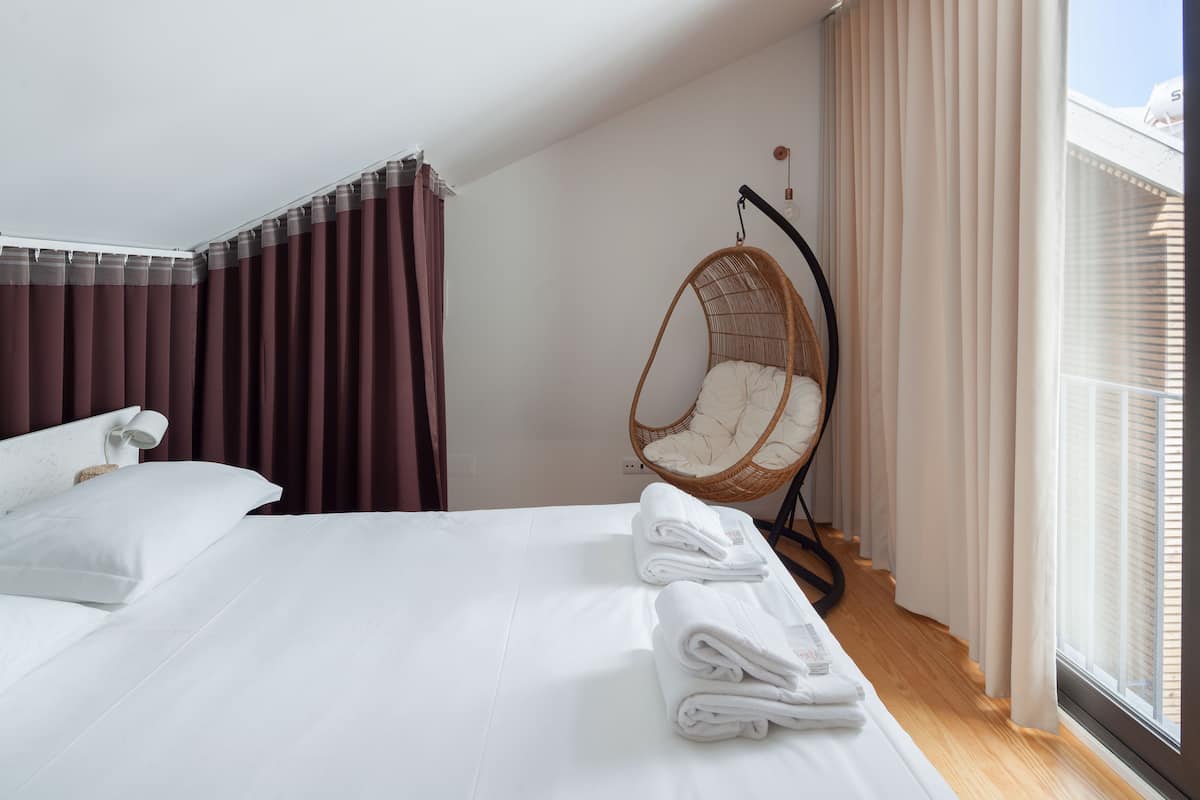 A modern hotel room with a large bed, a hanging chair, and a view of the sky through the window.