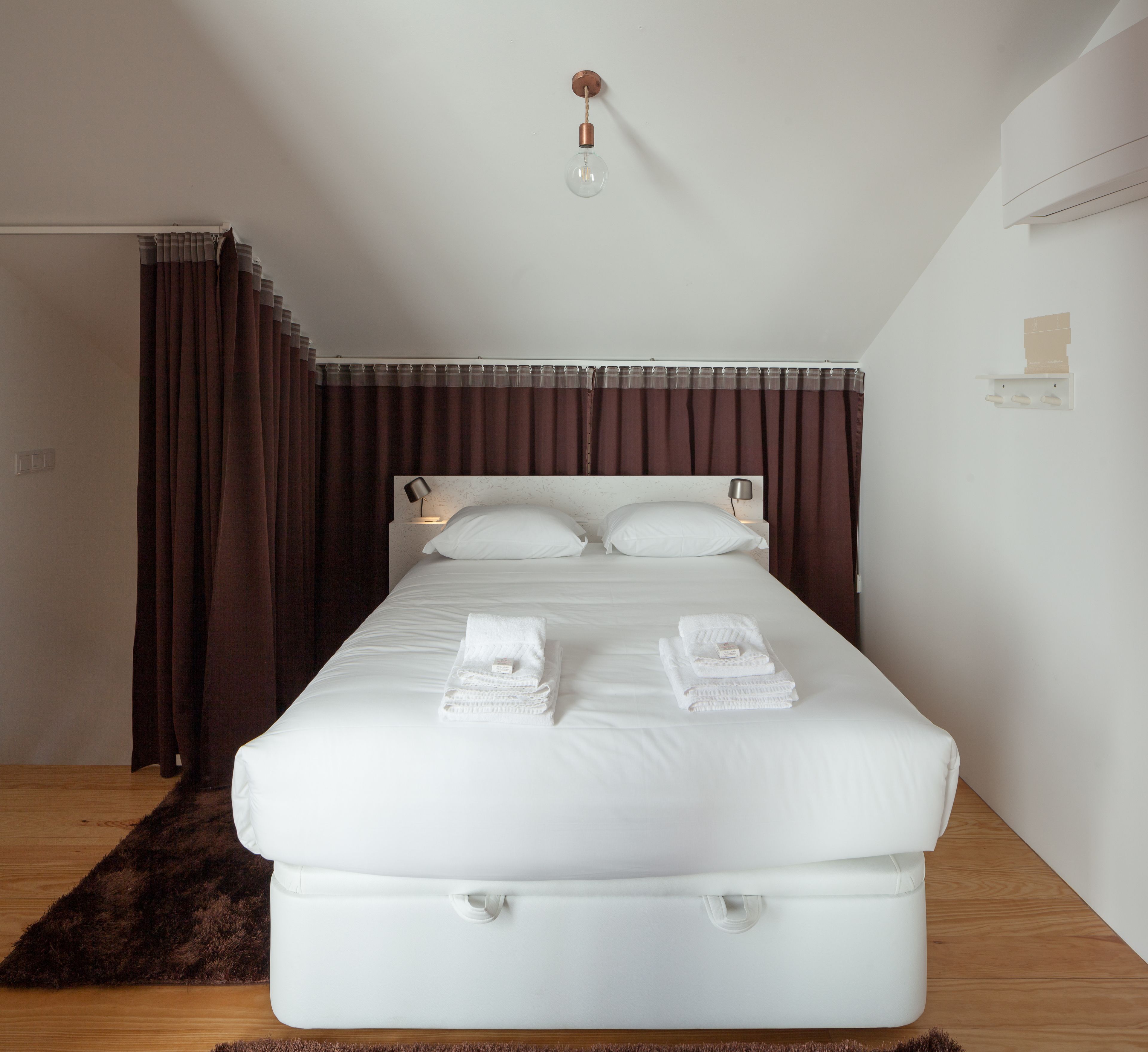 A neatly made bed with white linens and towels, a headboard with wall-mounted lamps, and a dark brown curtain.
