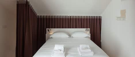 A neatly made bed with white linens and towels, a headboard with wall-mounted lamps, and a dark brown curtain.