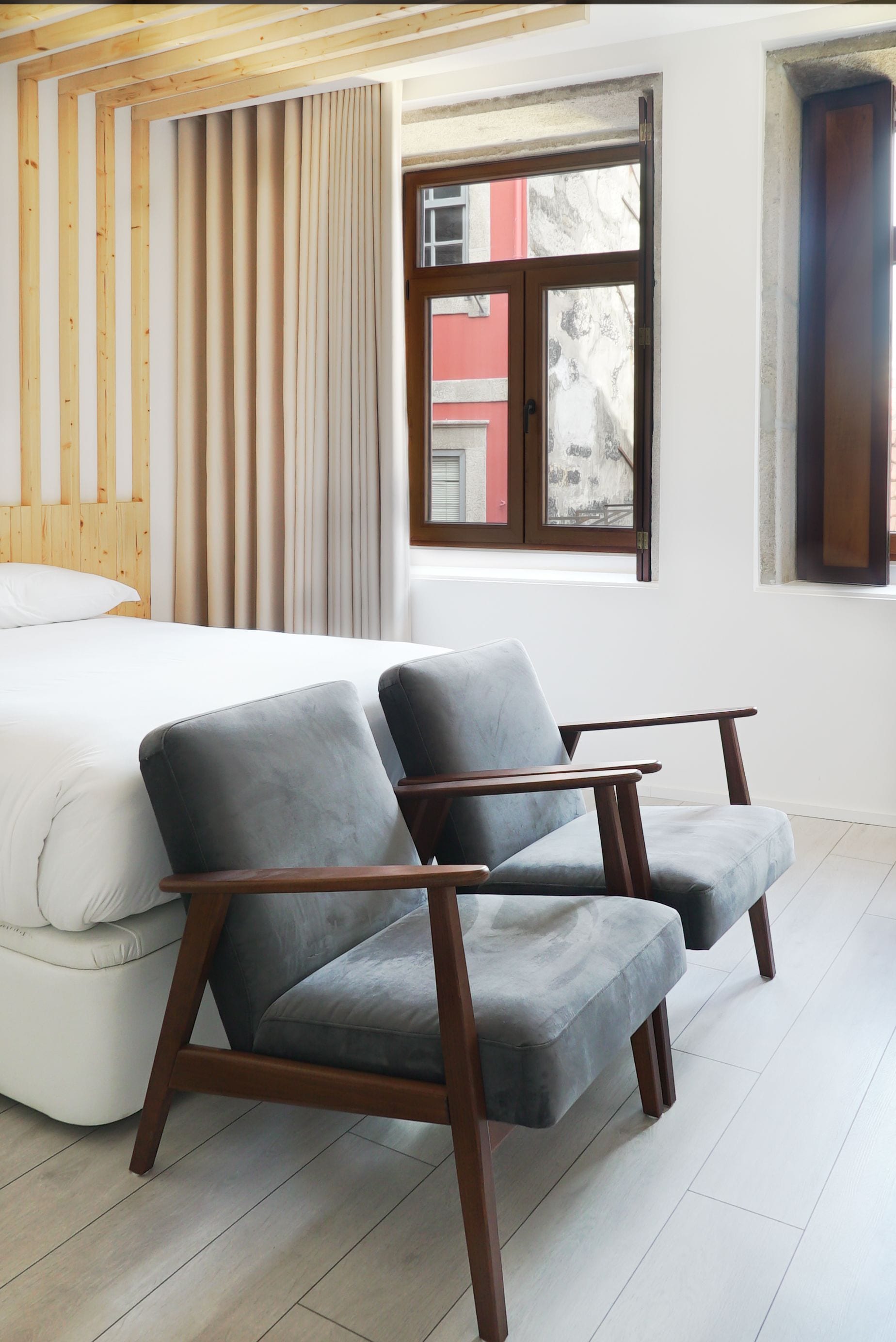 A bedroom with a bed, two grey armchairs, wooden headboard, and a window with a view of buildings.