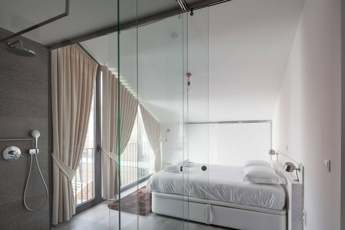 A modern bedroom with a glass partition, a shower, a bed, and a view of the outdoors.