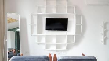 Quarto de hotel moderno com televisão central, prateleiras embutidas e área de estar com dois pufes.