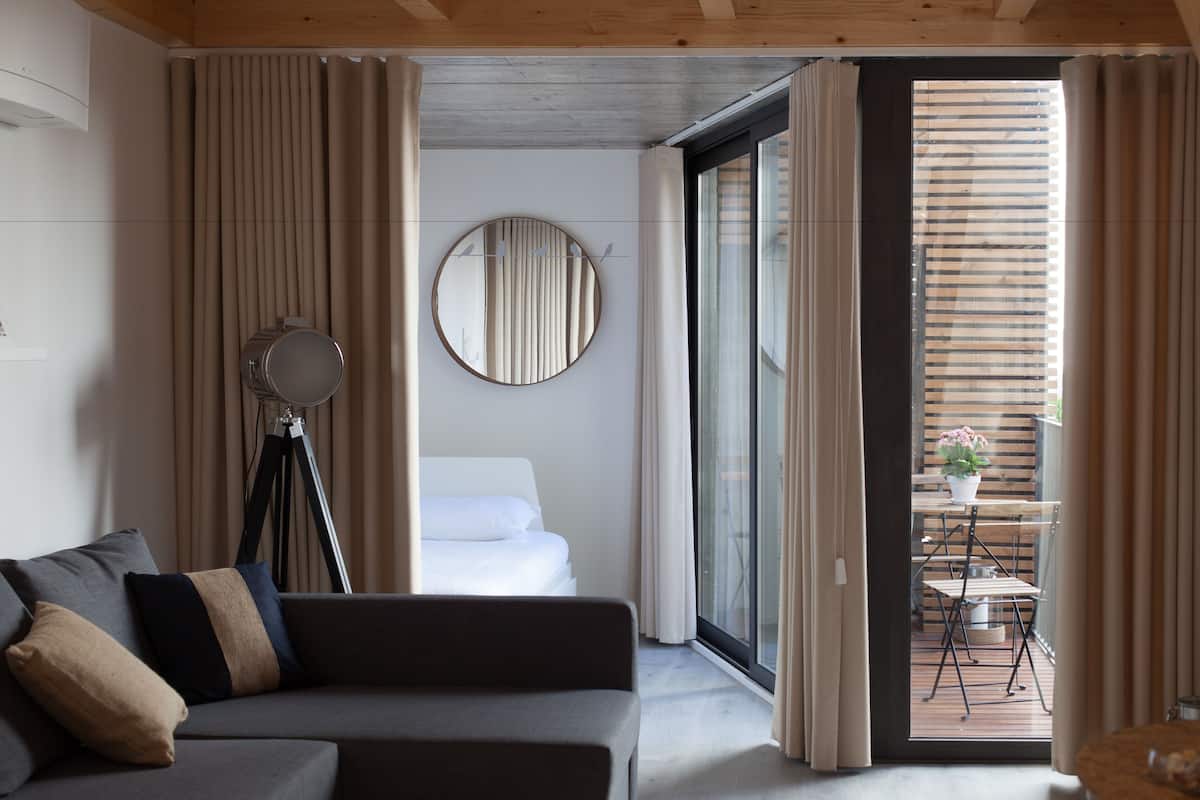 A modern hotel room with a grey sofa, a round mirror, a bed with white linens, and a sliding glass door leading to a balcony with a table and chairs.