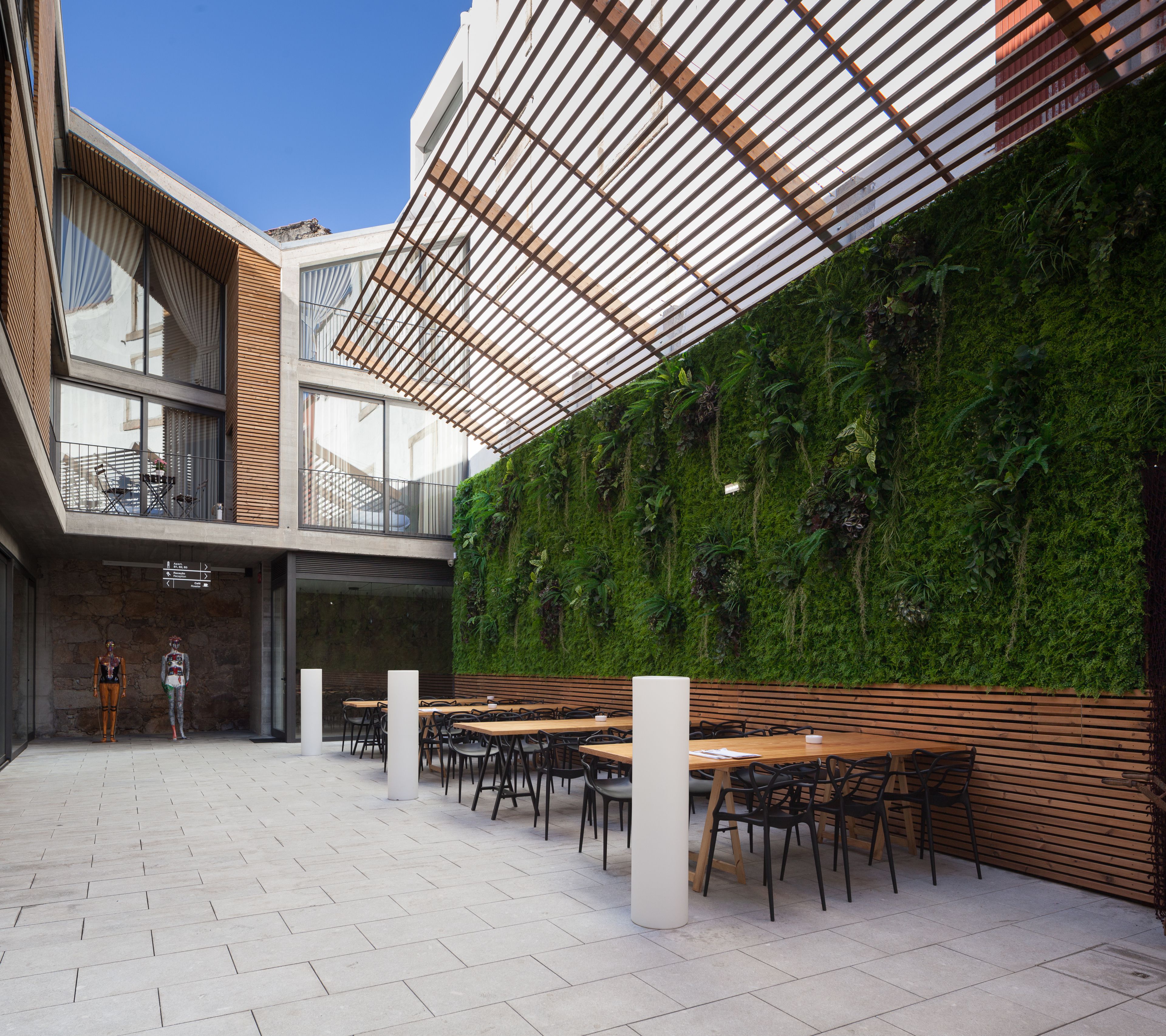 An outdoor dining area with wooden tables and chairs, a green vertical garden wall, and a modern building with large windows and a sloped roof.
