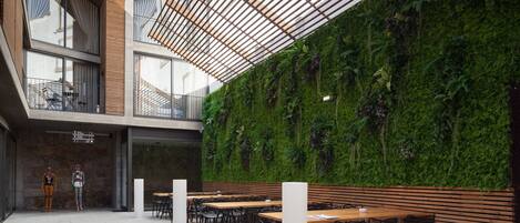 An outdoor dining area with wooden tables and chairs, a green vertical garden wall, and a modern building with large windows and a sloped roof.