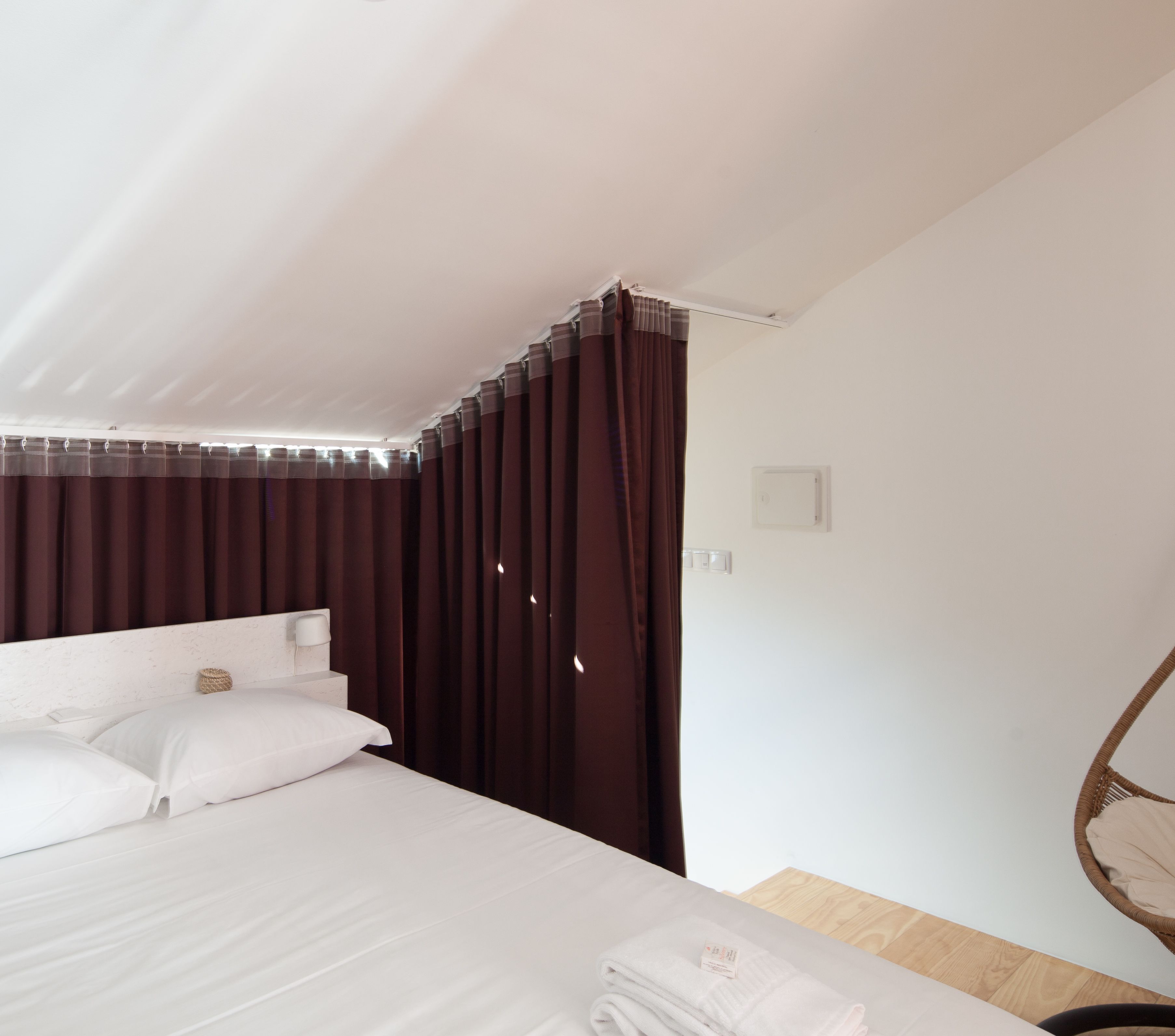 A bedroom with a bed, white bedding, a dark curtain, a bedside table, and a small wall-mounted light.