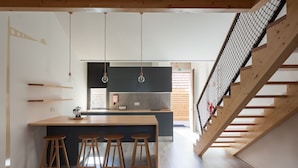 A modern kitchen with a central island, wooden stools, and a staircase with a wooden railing. - 7g Roaster Apartments (Vila Nova de Gaia)