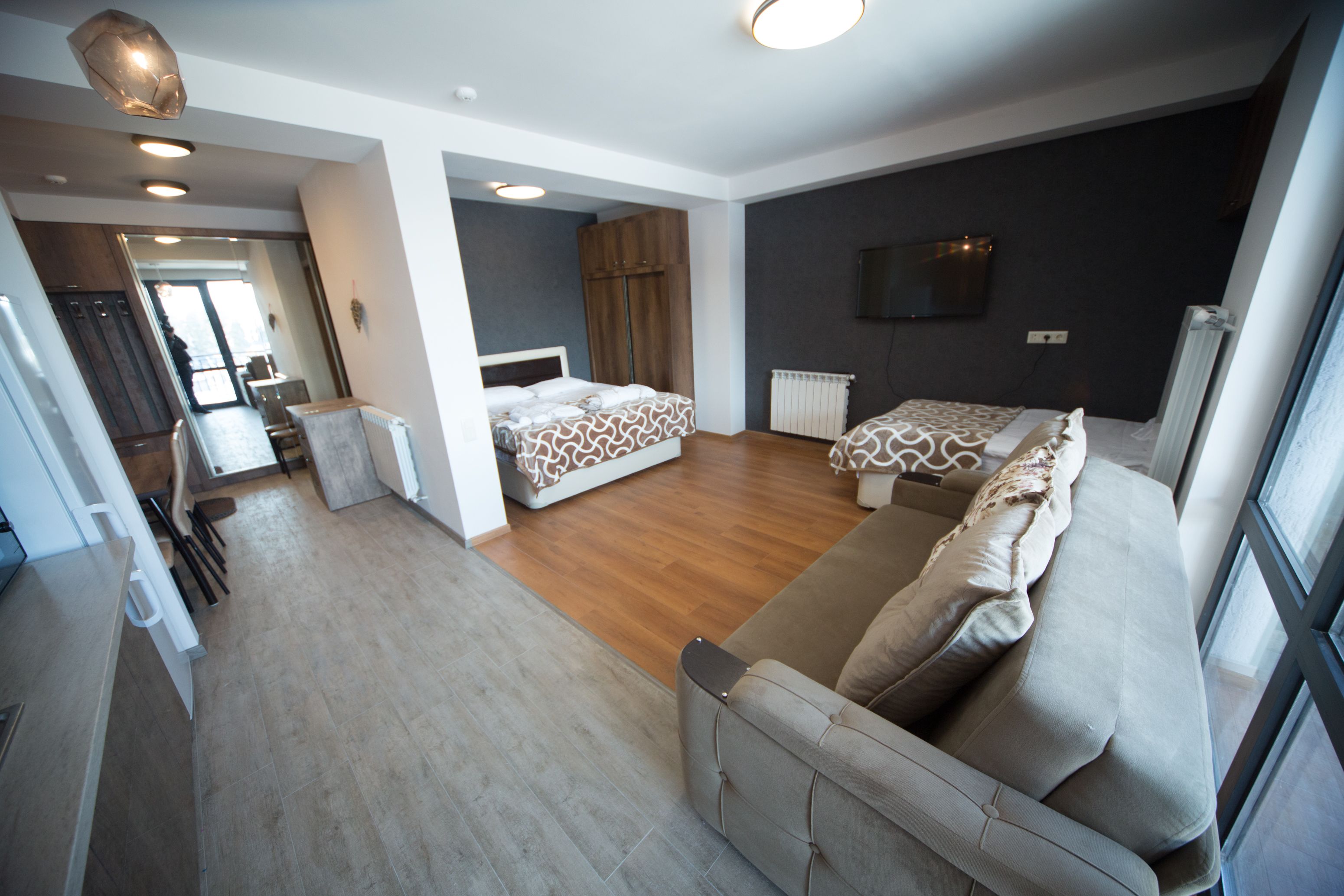 A modern hotel room with a sofa, two beds, wooden flooring, and a TV on the wall.