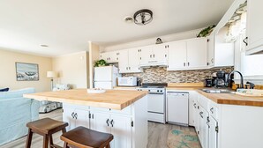 Moderni keittiö, jossa on valkoiset kaapit, puinen työtaso ja sisäänrakennettu uuni. Työtasolla on kaksi jakkaraa ja kahvinkeitin.