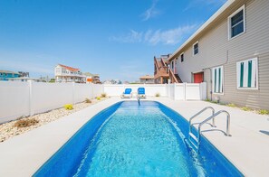 En swimmingpool med två blå solstolar, ett vitt staket och en byggnad med en röd dörr och gröna fönsterluckor.