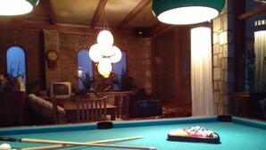 A pool table with billiard balls and cues, a clock, and a television in a room with brick walls and wooden beams. - Nicolas Village Club (Aigialeia)
