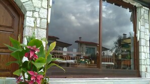 A stone building with a large window reflecting a house and a cloudy sky. - Nicolas Village Club (Aigialeia)