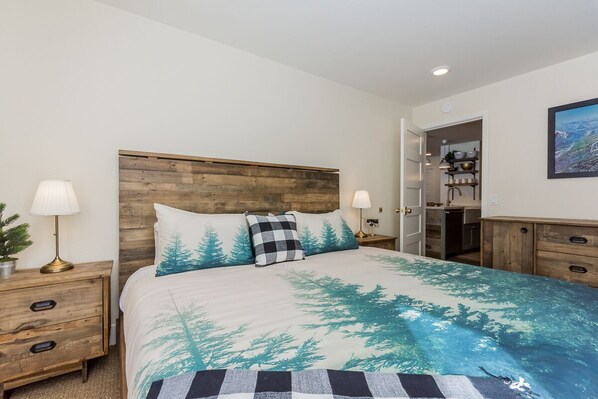 Un dormitorio con cabecera de madera, cama con edredón de motivos forestales, lámparas de noche, mesita de noche y vista hacia una zona de cocina.