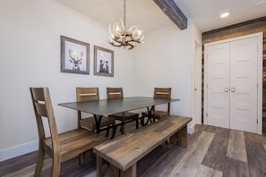 Et spiseområde med et bord med glasplade, træstole, en bænk og en lysekrone. Væggene er dekoreret med billeder i rammer, og i baggrunden ses et sæt dobbelte døre.
