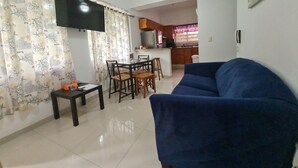 Una sala de estar con un sofá azul, una mesa de comedor con sillas, un televisor montado en la pared y un ventilador de techo.
