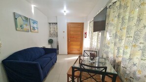 Una sala de estar con un sofá azul, una mesa de comedor de vidrio con sillas, una puerta de madera y un televisor en la pared.