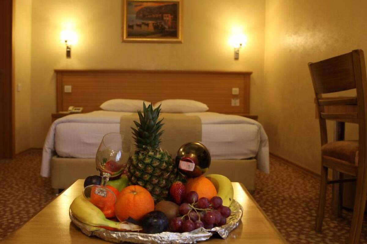 A hotel room with a bed, a table with fruit and a glass of wine, and a painting on the wall.