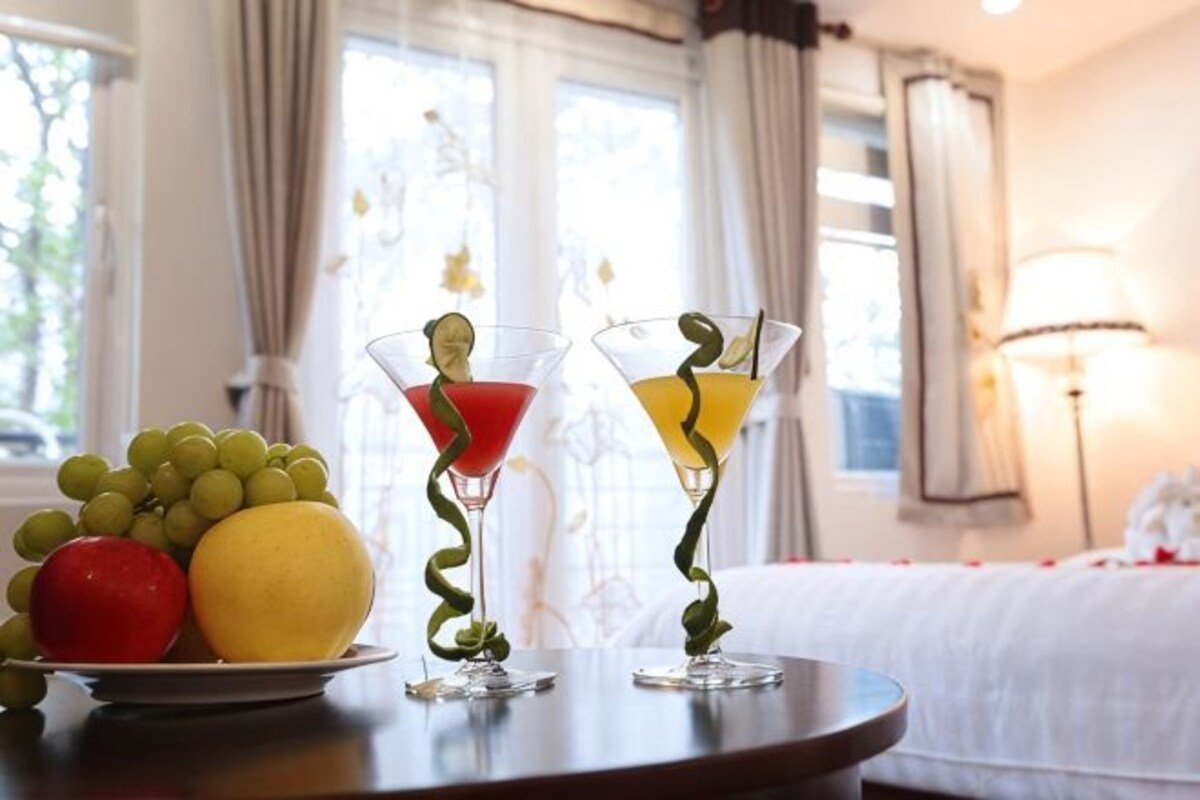 Une table avec deux verres à martini, une assiette de fruits et une lampe, dans une pièce où se trouvent un lit et des rideaux.