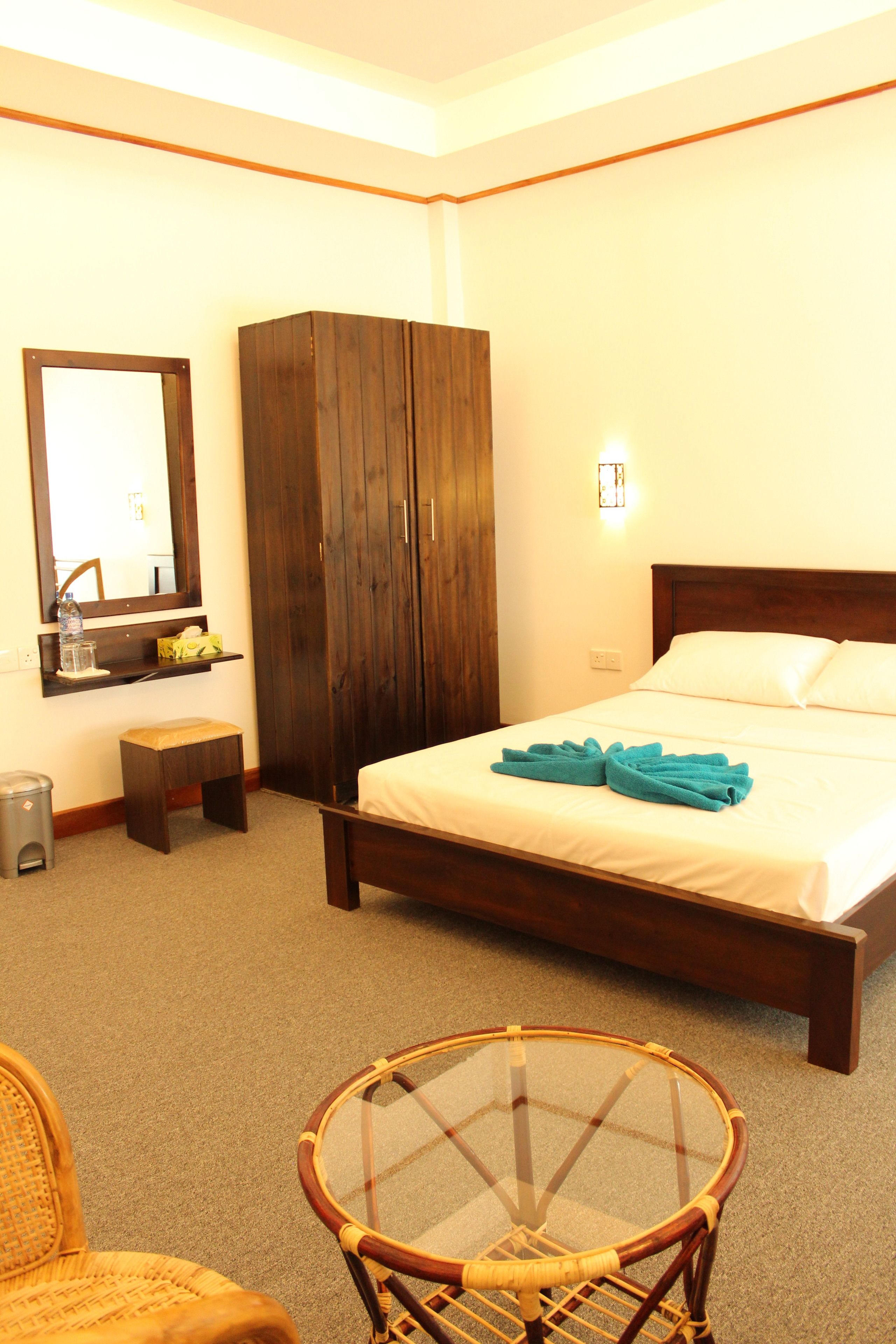 A hotel room with a bed, a wooden wardrobe, a small stool, a wicker chair, and a glass table.