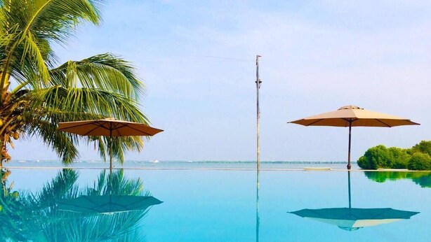 Una piscina infinita azul cristalina con dos sombrillas, palmeras y vista al océano.
