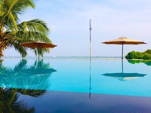 Une magnifique piscine à débordement d’un bleu profond, agrémentée de deux parasols, de palmiers et offrant une vue sur l’océan.