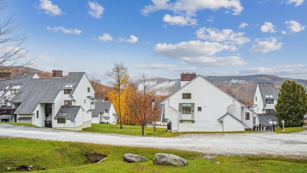 Une rangée de maisons blanches aux toits inclinés, une zone herbeuse au premier plan et un paysage montagneux en arrière-plan, le tout sous un ciel partiellement nuageux.