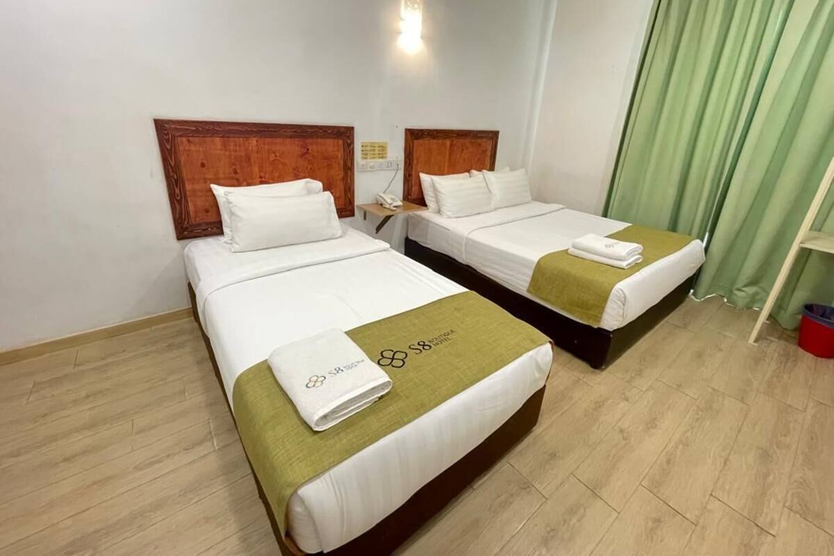 Habitación de hotel con dos camas individuales, cada una con cabecero de madera, ropa de cama blanca y una toalla verde. Hay una mesita de noche con un teléfono y una lámpara. La habitación tiene piso de madera y una cortina verde en la ventana.