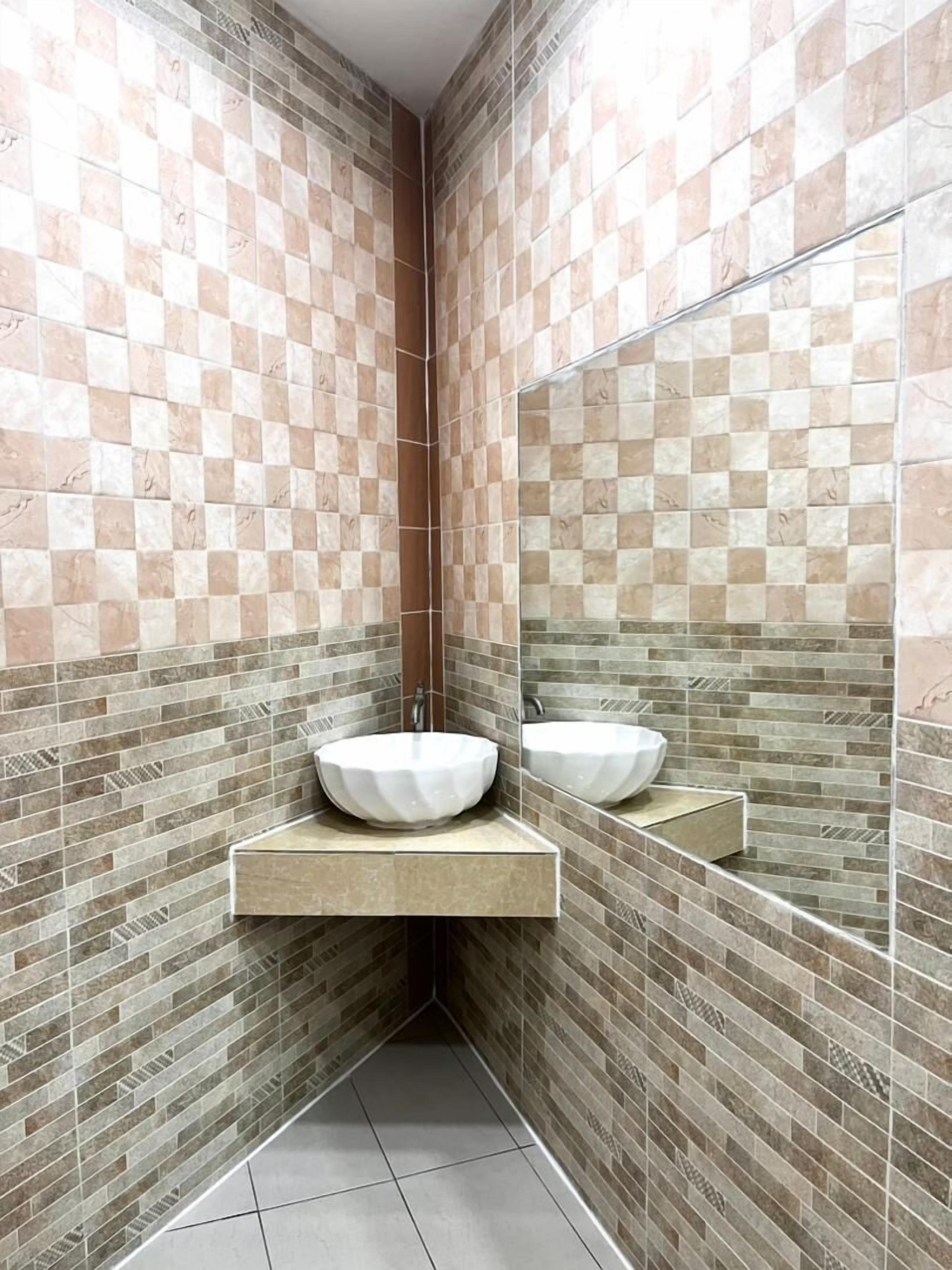 Un baño con pared de baldosas a cuadros, un lavamanos en esquina con espejo y suelo de baldosas.