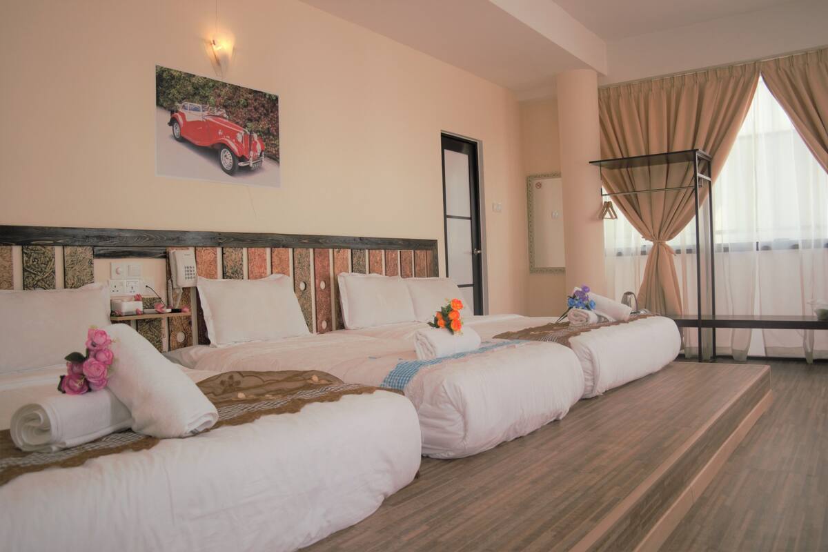 Habitación de hotel con dos camas, cabecera de madera, un cuadro de un coche, una ventana con cortinas y una mesita de noche con una lámpara.