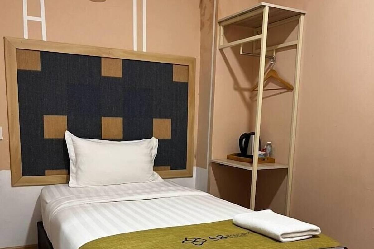 Una cama bien hecha con colcha blanca y verde, un cabecero de madera y una mesita de noche con una taza y una botella.