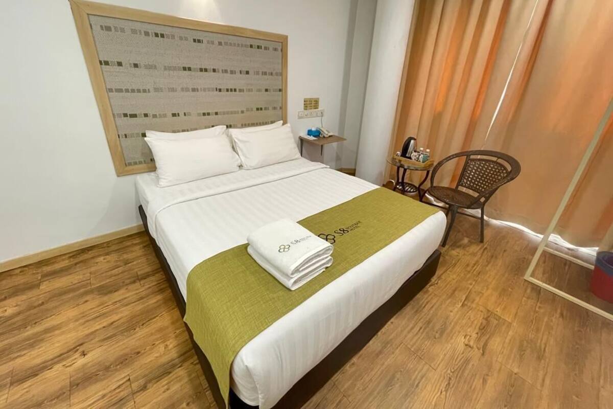 Habitación de hotel con una cama grande, piso de madera, una manta verde, una silla y una lámpara fijada a la pared.