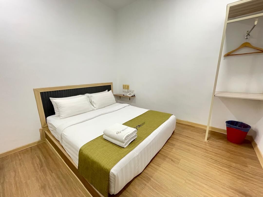 Una cama bien tendida con una manta verde, una almohada blanca y una toalla doblada al pie. Junto a la cama, una mesita con un teléfono y un bloc de notas. Se ve un cubo de basura rojo en una esquina.