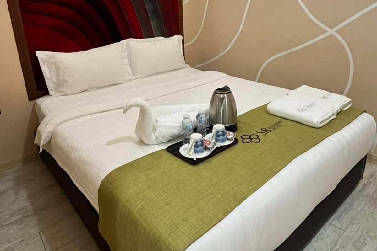 Una habitación de hotel con una cama, un plaid verde, una bandeja con una tetera y un cabecero rojo.