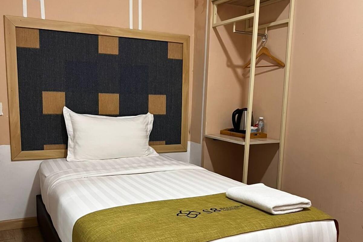 Una cama bien hecha con colcha blanca y verde, cabecera de madera y una pequeña estantería con un hervidor y una botella.