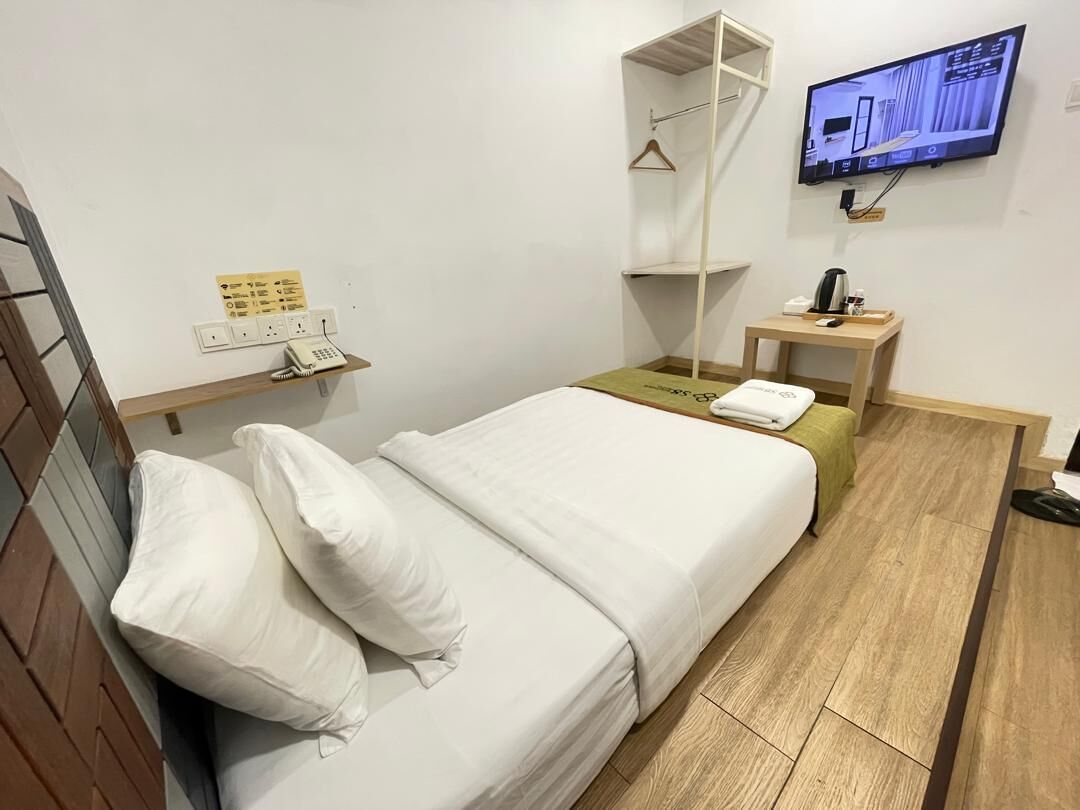 A hotel room with a bed, a TV mounted on the wall, a bedside table with a phone and a kettle, and a wooden floor.
