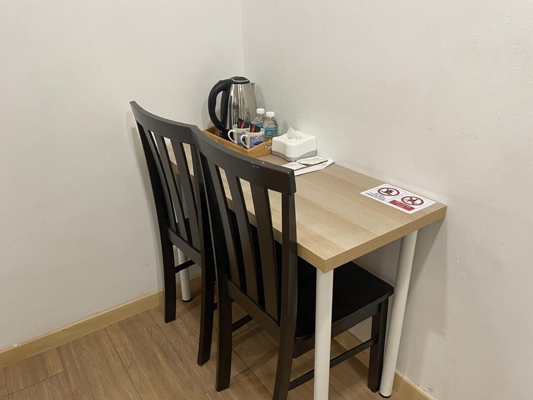Une pièce d’angle avec une table en bois, deux chaises noires, une bouilloire, une boîte de mouchoirs et une bouteille d’eau.