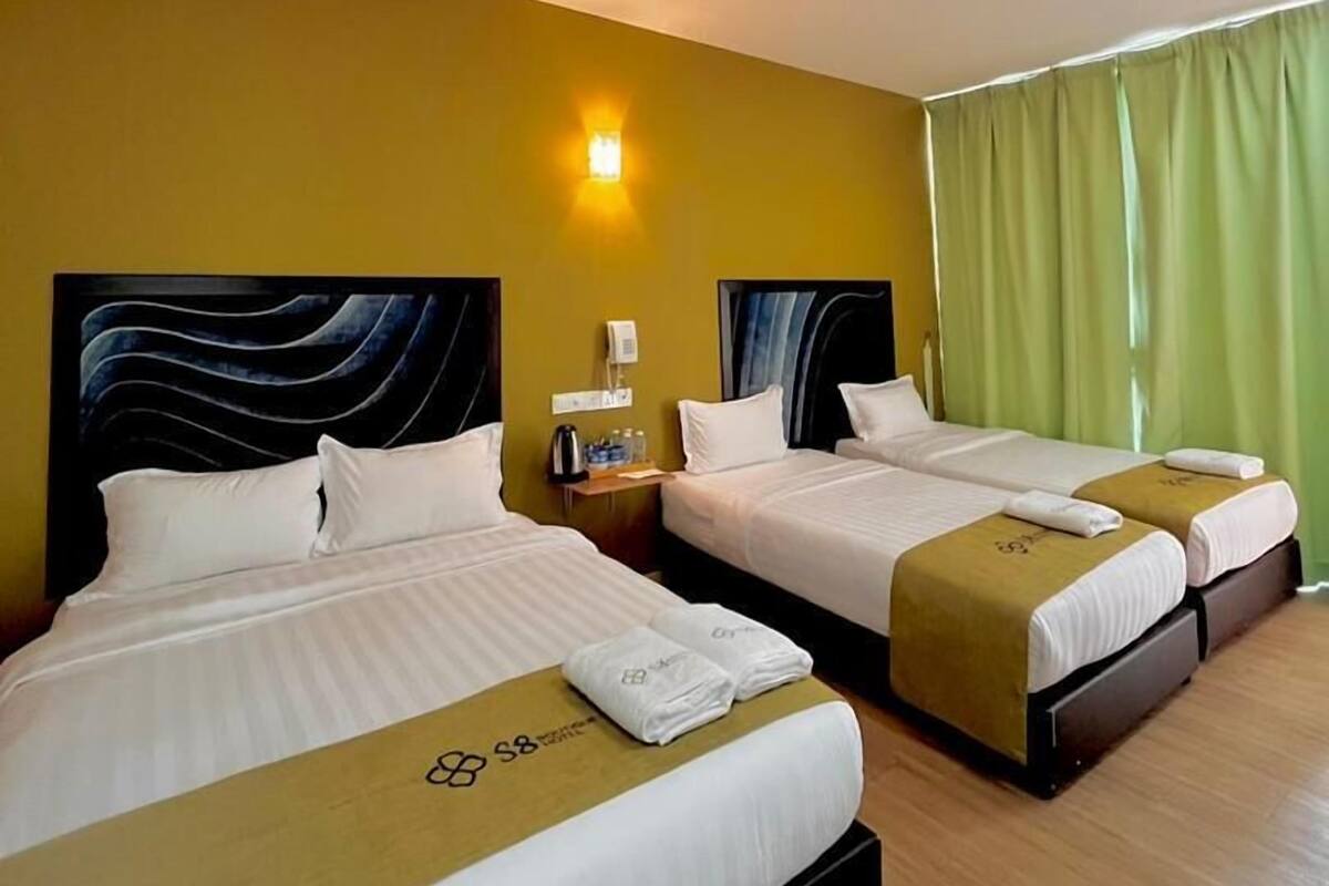 Habitación de hotel con dos camas, una mesita de noche, una lámpara fijada a la pared y una cortina verde.