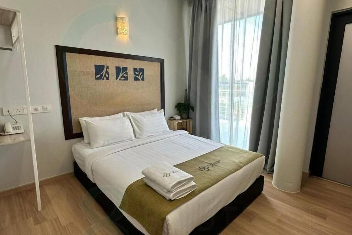 Una cama bien tendida con sábanas blancas y un plaid verde, un aplique de pared y una ventana con cortinas traslúcidas.