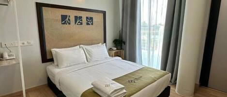 Cama bem arrumada com lençóis brancos e manta verde, luminária fixada na parede e janela com cortinas leves.