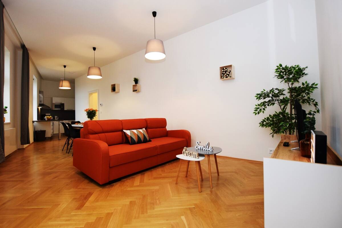 Uma sala de estar moderna com um sofá vermelho, piso de madeira, luminárias pendentes e uma pequena mesa com uma placa.