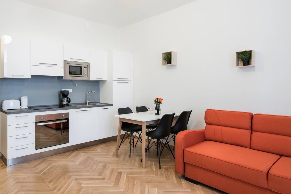 Cozinha moderna com armários brancos, micro-ondas e máquina de café. Há uma mesa de jantar com cadeiras e um sofá vermelho.
