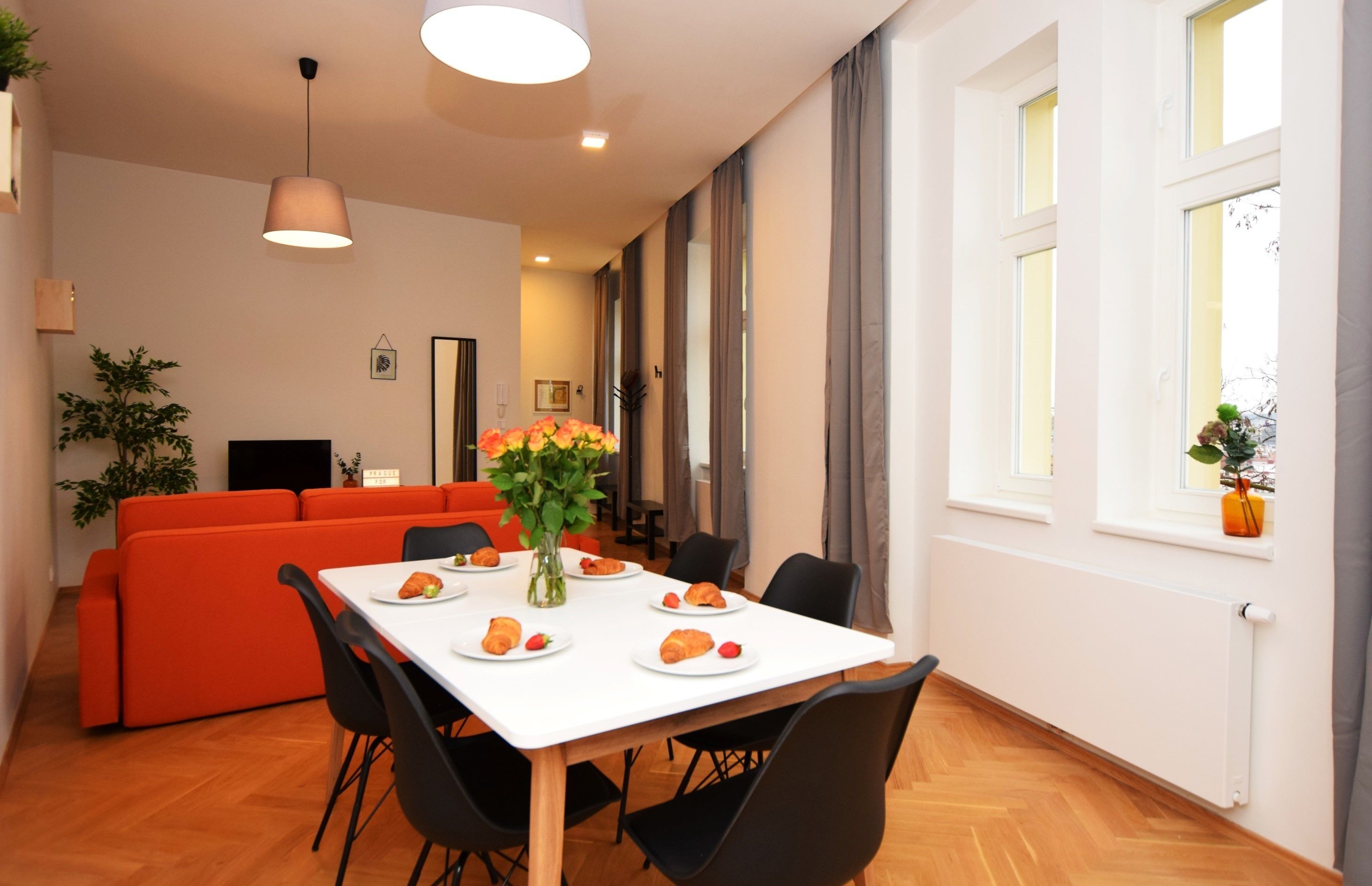 A dining area with a white table, black chairs, and a vase with flowers. There is a red sofa and a plant in the background.