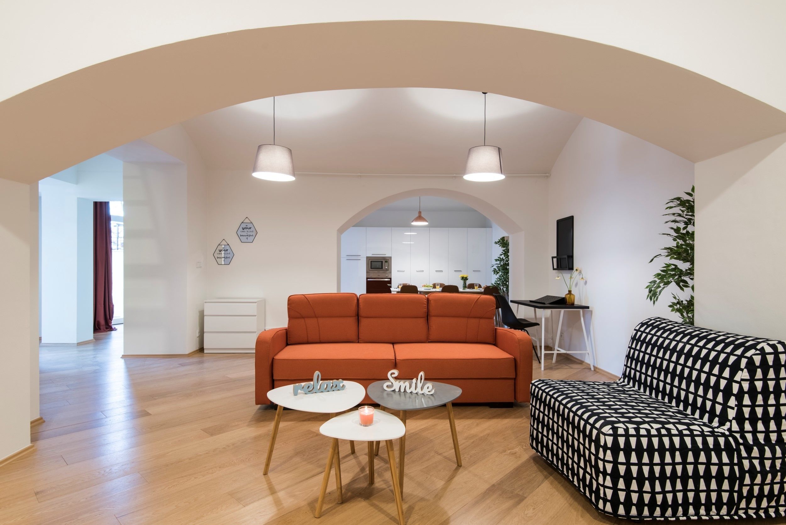 A modern living room with an orange sofa, a black and white patterned armchair, and two small white tables with decorative items.