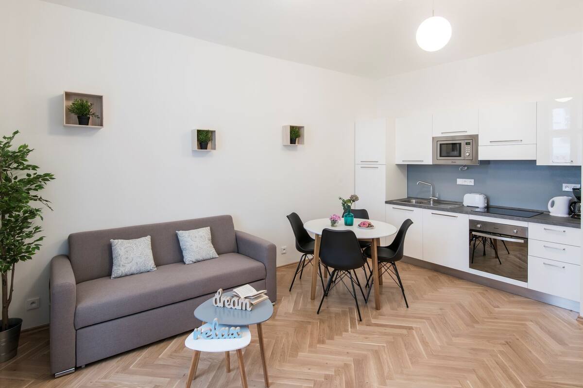 Um apartamento moderno com sofá cinzento, uma pequena mesa redonda, uma mesa de jantar com cadeiras, cozinha com armários brancos e um micro-ondas, e uma planta em vaso.