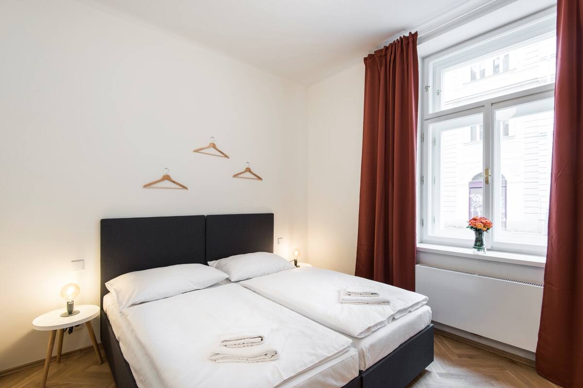 Cama de casal com roupa de cama branca, mesa de cabeceira com abajur e janela com cortinas.
