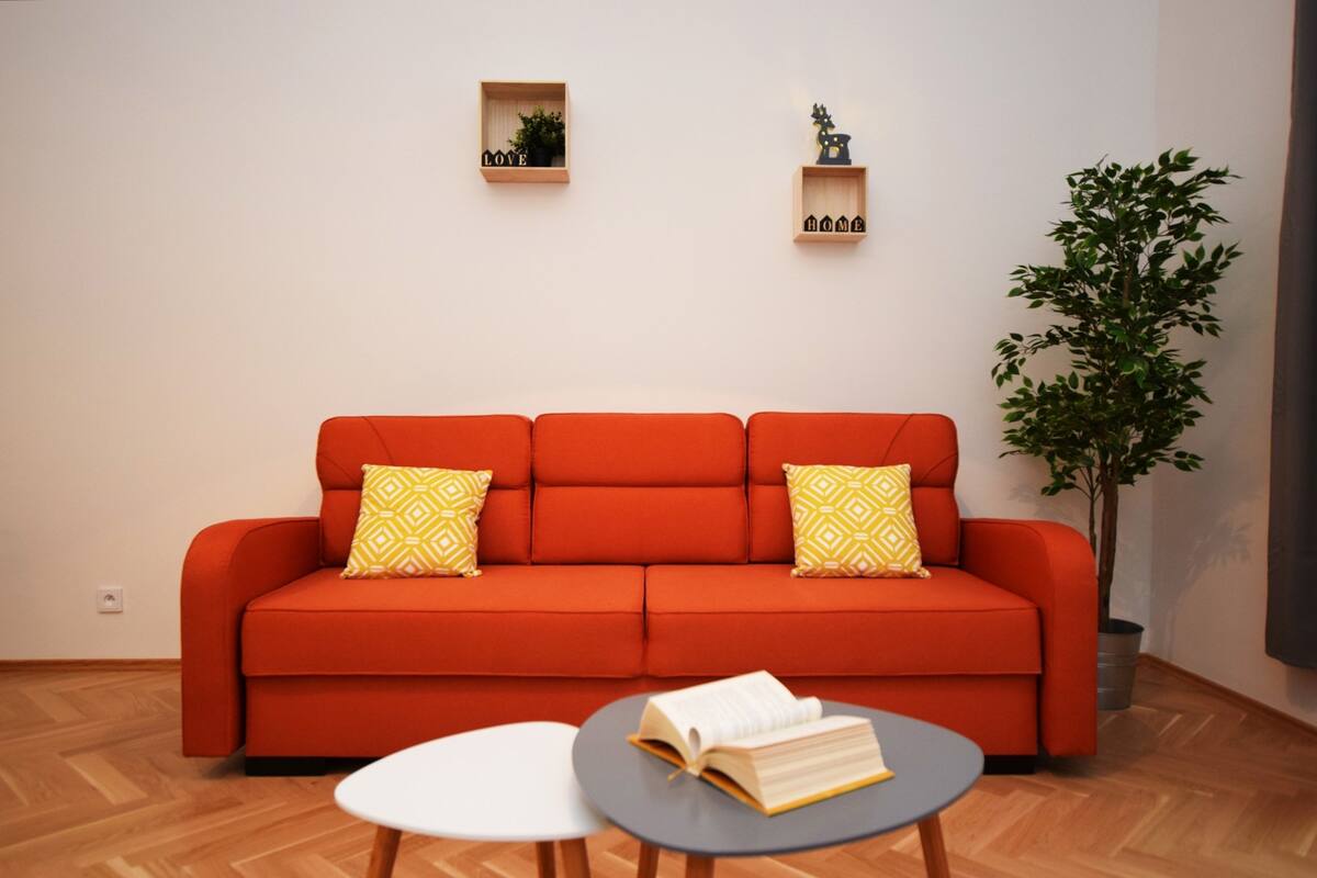 Uma sala de estar moderna com um sofá vermelho, duas almofadas estampadas em amarelo, uma mesinha com um livro e uma planta em vaso.