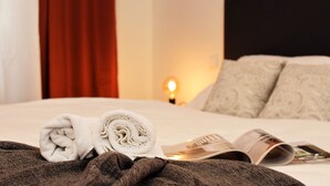 Una cama bien tendida con una toalla enrollada y una revista, una lámpara de noche y un cojín estampado.