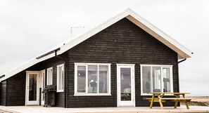 A black wooden building with white-framed windows, a door, and a bench on the deck. - Gullfoss & Geysir Luxury Cabin (Bláskógabyggd)